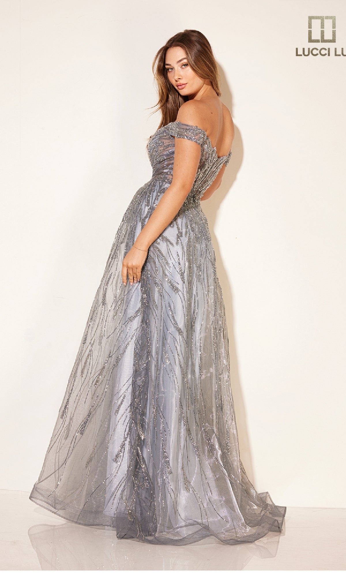 A woman in the Lucci Lu C8044 pastel blue prom ball gown, featuring an off-the-shoulder neckline, fitted sheer corset bodice, and flowing floor-length skirt, stands against a plain light background.